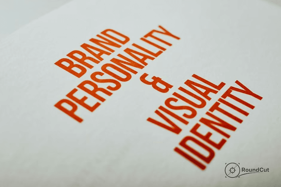 A close-up, angled shot of white textured paper displaying the orange-red text "BRAND PERSONALITY & VISUAL IDENTITY